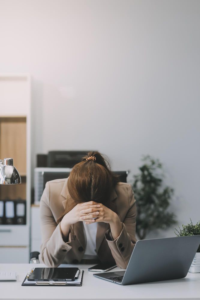 A woman sits at her desk, resting her head in her hands, appearing stressed or overwhelmed.